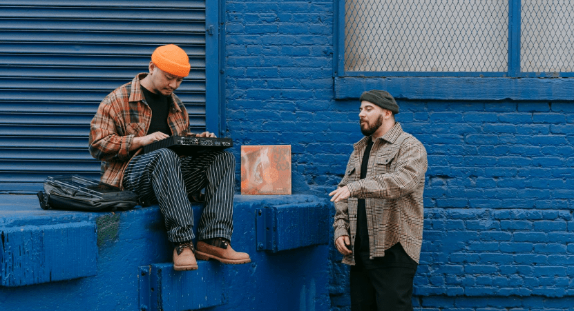 Two male creatives sitting against a bright blue brick wall using a music mixer.