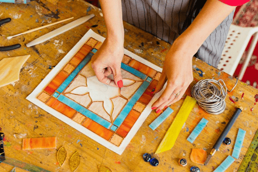 Close-up of hands working on a colorful mosaic tile artwork on a wooden table.
