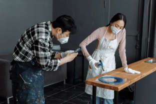 Two creatives wearing aprons and masks working together on a resin art project in a studio.